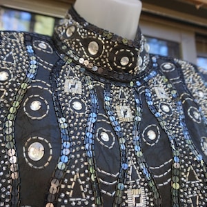 May include: Black, long-sleeved top with intricate sequin and bead embellishments. The high collar and shoulders are heavily decorated with silver sequins and clear, round rhinestones. The design features geometric patterns and a touch of iridescent blue.