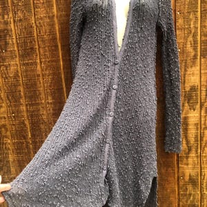 K&ouml;nnte beinhalten: Lange, gekn&ouml;pfte Strickjacke aus strukturiertem, grauem Strickstoff. Die Strickjacke hat lange &Auml;rmel, einen V-Ausschnitt und einen geschlitzten Saum. Der Stoff hat eine noppige Textur und die Strickjacke scheint leicht zu sein.
