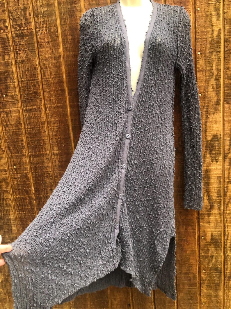 K&ouml;nnte beinhalten: Lange, gekn&ouml;pfte Strickjacke aus strukturiertem, grauem Strickstoff. Die Strickjacke hat lange &Auml;rmel, einen V-Ausschnitt und einen geschlitzten Saum. Der Stoff hat eine noppige Textur und die Strickjacke scheint leicht zu sein.
