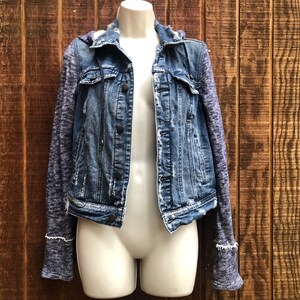 May include: A blue denim jacket with a hood and knit sleeves. The jacket has a button-up front, two chest pockets, and a distressed look. The sleeves are a heathered blue-gray knit material, and the jacket is displayed on a mannequin.