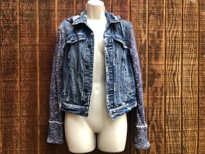May include: A blue denim jacket with a hood and knit sleeves. The jacket has a button-up front, two chest pockets, and a distressed look. The sleeves are a heathered blue-gray knit material, and the jacket is displayed on a mannequin.
