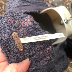 K&ouml;nnte beinhalten: Nahaufnahme eines dunkelgrauen Strickkleidungsst&uuml;cks mit einem strukturierten, offen gewebten Design. Ein kleines, kupferfarbenes Etikett mit der Aufschrift "Free People" ist angebracht. Ein wei&szlig;es Etikett mit Text in Franz&ouml;sisch und Englisch ist ebenfalls sichtbar.