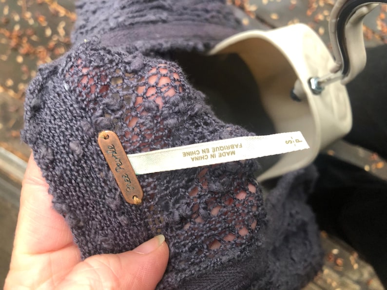 K&ouml;nnte beinhalten: Nahaufnahme eines dunkelgrauen Strickkleidungsst&uuml;cks mit einem strukturierten, offen gewebten Design. Ein kleines, kupferfarbenes Etikett mit der Aufschrift "Free People" ist angebracht. Ein wei&szlig;es Etikett mit Text in Franz&ouml;sisch und Englisch ist ebenfalls sichtbar.