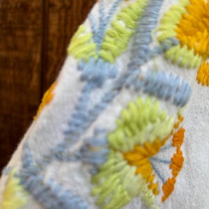 May include: Close-up of a white fabric with embroidered floral design. The embroidery features yellow, light blue, and green colors. The stitching is detailed, creating a textured appearance. The design is likely part of a garment.