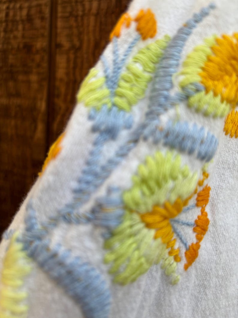 May include: Close-up of a white fabric with embroidered floral design. The embroidery features yellow, light blue, and green colors. The stitching is detailed, creating a textured appearance. The design is likely part of a garment.