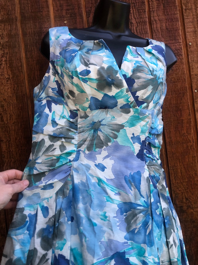May include: Sleeveless dress with a floral pattern in shades of blue, gray, and white. The dress features a V-neckline and a fitted bodice with pleating details. The skirt is flared, and the fabric appears to be lightweight.