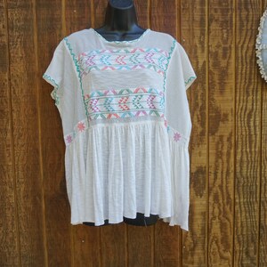 May include: White embroidered tunic top with colorful geometric patterns across the chest and sleeves. The top has a loose, flowing fit with short sleeves and a gathered waist. The embroidery features pink, teal, and green accents. The background is a wooden wall.