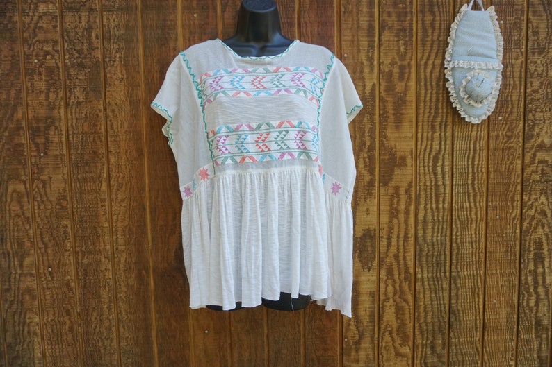 May include: White embroidered tunic top with colorful geometric patterns across the chest and sleeves. The top has a loose, flowing fit with short sleeves and a gathered waist. The embroidery features pink, teal, and green accents. The background is a wooden wall.
