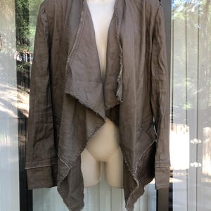 K&ouml;nnte beinhalten: Offene Jacke in Braun mit drapiertem Design. Die Jacke hat lange &Auml;rmel und rohe, ausgefranste Kanten an Revers und Saum. Der Stoff sieht nach leichtem Leinen oder einem &auml;hnlichen Material aus. Die Jacke wird auf einer Schaufensterpuppe pr&auml;sentiert.