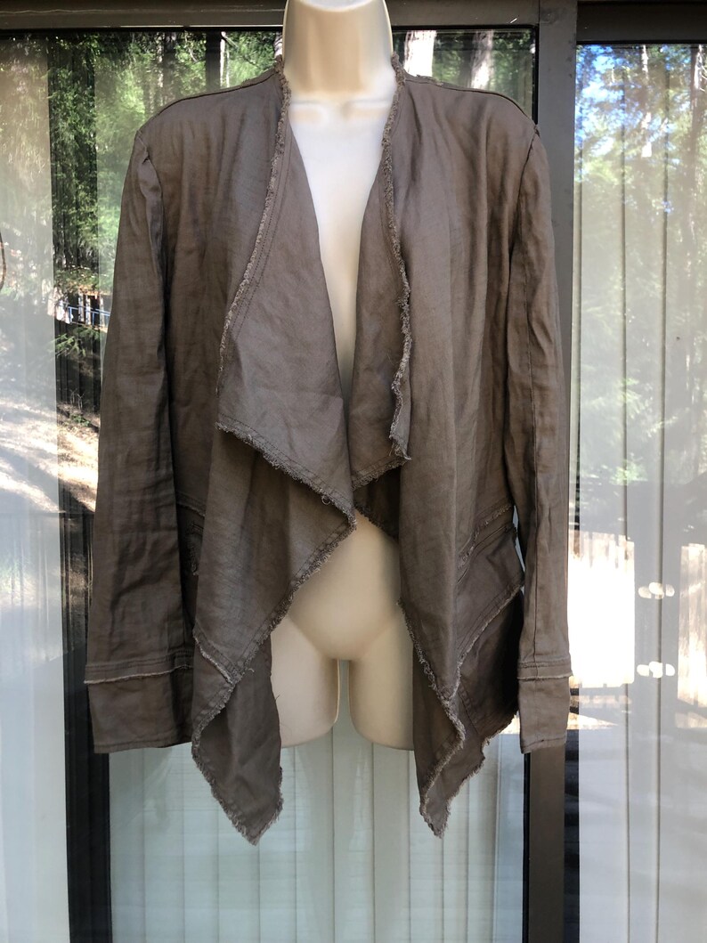 K&ouml;nnte beinhalten: Offene Jacke in Braun mit drapiertem Design. Die Jacke hat lange &Auml;rmel und rohe, ausgefranste Kanten an Revers und Saum. Der Stoff sieht nach leichtem Leinen oder einem &auml;hnlichen Material aus. Die Jacke wird auf einer Schaufensterpuppe pr&auml;sentiert.
