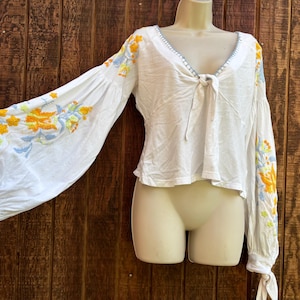 May include: White blouse with long, voluminous sleeves and a V-neckline. The sleeves and neckline are adorned with colorful floral embroidery in shades of yellow, orange, and blue. The blouse has a tie detail at the neckline and tie cuffs.