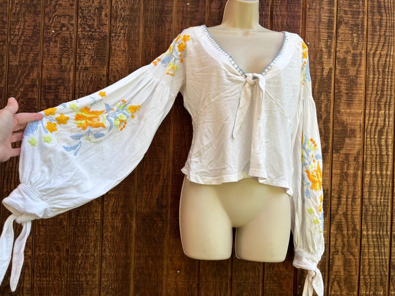 May include: White blouse with long, voluminous sleeves and a V-neckline. The sleeves and neckline are adorned with colorful floral embroidery in shades of yellow, orange, and blue. The blouse has a tie detail at the neckline and tie cuffs.