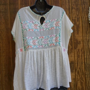 May include: White, short-sleeved tunic top with colorful embroidered geometric patterns across the chest and neckline. The top has a keyhole neckline and a gathered, flowing skirt. The embroidery includes pink, blue, and green accents.