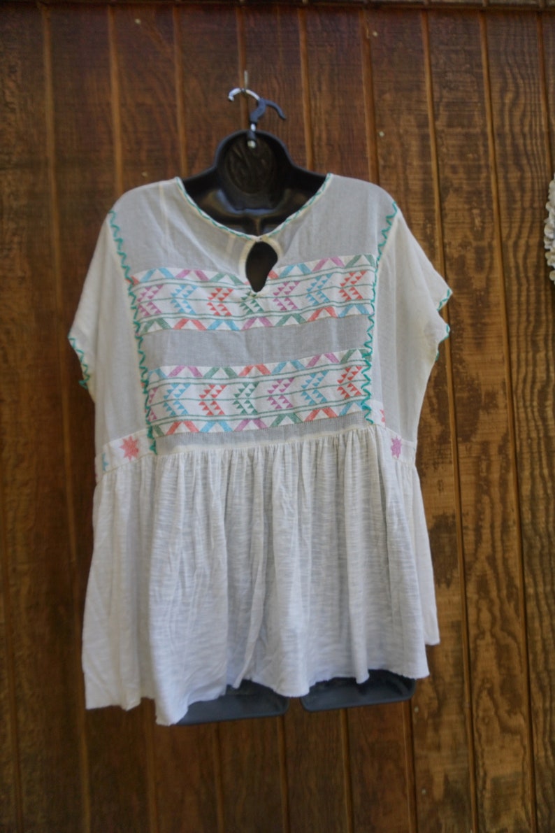 May include: White, short-sleeved tunic top with colorful embroidered geometric patterns across the chest and neckline. The top has a keyhole neckline and a gathered, flowing skirt. The embroidery includes pink, blue, and green accents.
