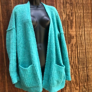 May include: A teal blue open-front cardigan with long sleeves and two pockets. The cardigan is made of a soft, fuzzy material.