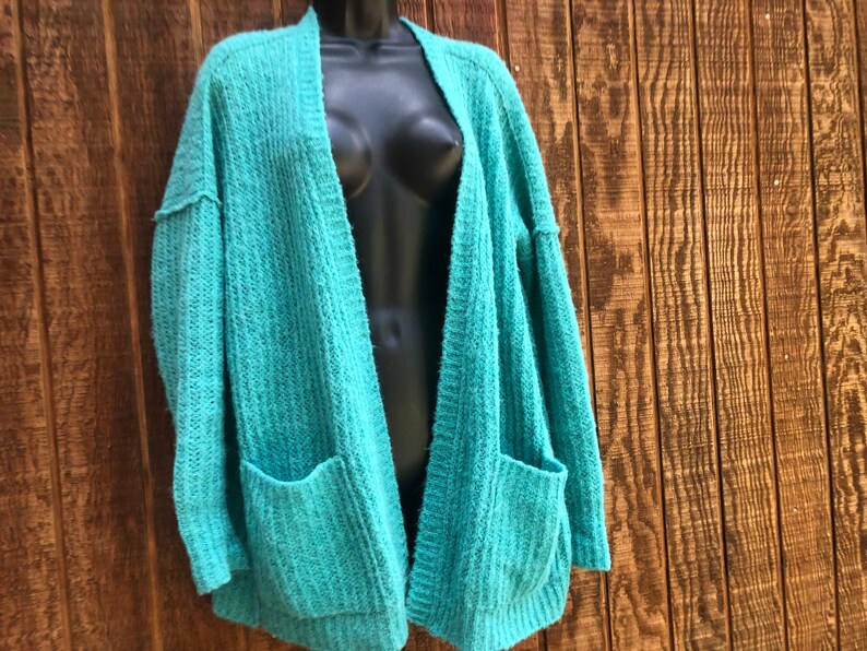 May include: A teal blue open-front cardigan with long sleeves and two pockets. The cardigan is made of a soft, fuzzy material.