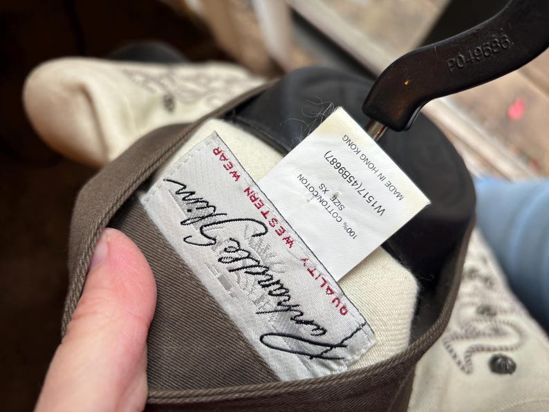 Puede incluir: Primer plano de una prenda Western wear marr&oacute;n y beige. La etiqueta dice "Vanhandle Glim QUALITY WESTERN WEAR". Una etiqueta indica que la prenda es XS, 100% algod&oacute;n y fabricada en Hong Kong.