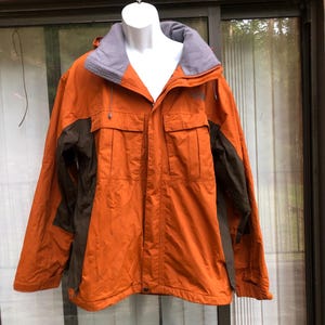 May include: An orange and brown jacket with a gray collar and hood. The jacket has two front pockets and a zipper closure. The sleeves are brown, and the jacket is hanging on a white mannequin.