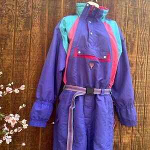 May include: A purple one-piece ski suit featuring teal and pink color-blocked panels, a high collar, a front placket with snap buttons, a chest pocket, and a matching adjustable belt with a black plastic buckle. A small triangular patch reads 7.