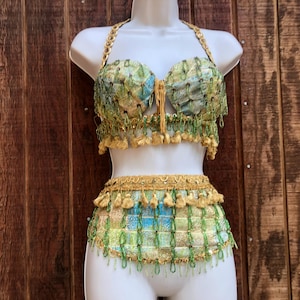 May include: A two-piece costume with a halter top and a skirt. The top and skirt are decorated with green, gold, and blue accents. The halter top has a gold chain strap. The skirt has a fringe of gold tassels.