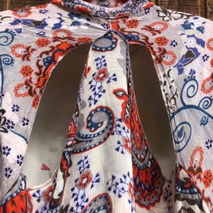 May include: A patterned, open-back top in cream, orange, blue, and gray. The fabric features a paisley and floral design. The top is hanging on a white hanger against a wooden background.