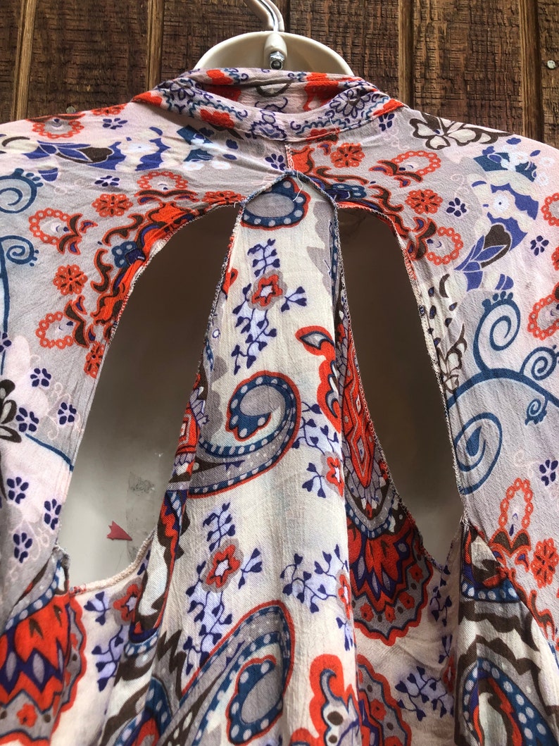 May include: A patterned, open-back top in cream, orange, blue, and gray. The fabric features a paisley and floral design. The top is hanging on a white hanger against a wooden background.