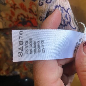May include: A white fabric tag with text indicating the garment is made of 100% rayon. The tag also includes care symbols and the text "MADE IN INDIA". The background shows a patterned fabric with red, blue, and beige colors.