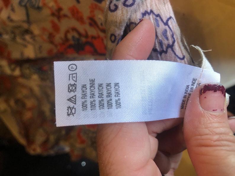 May include: A white fabric tag with text indicating the garment is made of 100% rayon. The tag also includes care symbols and the text "MADE IN INDIA". The background shows a patterned fabric with red, blue, and beige colors.