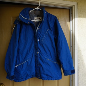 May include: A vibrant blue jacket with a hood, hanging on a white hanger. The jacket features a zipper closure, multiple pockets, and a logo on the chest. The jacket is made of a durable material, suitable for outdoor activities.