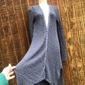 K&ouml;nnte beinhalten: Langer Cardigan mit Kn&ouml;pfen aus strukturiertem, dunkelblauem Stoff. Der Cardigan hat lange &Auml;rmel und einen asymmetrischen Saum. Das Kleidungsst&uuml;ck wird auf einer Schaufensterpuppe vor einem Holzgrund gezeigt.