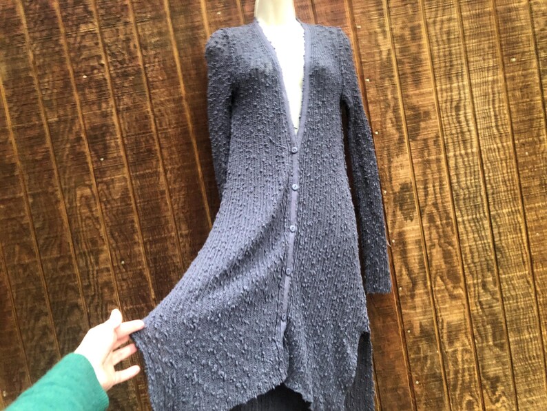 K&ouml;nnte beinhalten: Langer Cardigan mit Kn&ouml;pfen aus strukturiertem, dunkelblauem Stoff. Der Cardigan hat lange &Auml;rmel und einen asymmetrischen Saum. Das Kleidungsst&uuml;ck wird auf einer Schaufensterpuppe vor einem Holzgrund gezeigt.