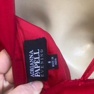 K&ouml;nnte beinhalten: Nahaufnahme eines roten Abendkleides mit Spaghettitr&auml;gern. Das schwarze Etikett lautet "Adrianna Papell Evening" und "Made in India". Das Kleid ist Gr&ouml;&szlig;e 6.