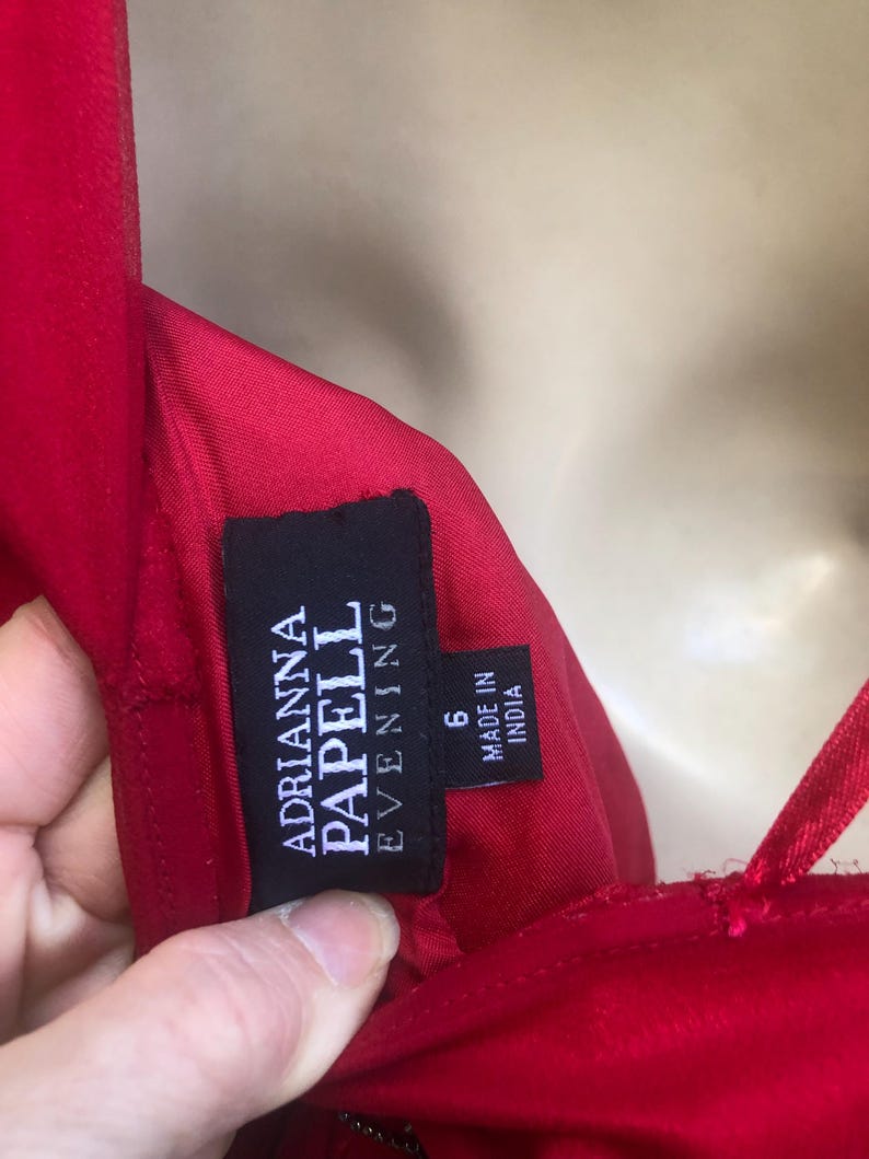 K&ouml;nnte beinhalten: Nahaufnahme eines roten Abendkleides mit Spaghettitr&auml;gern. Das schwarze Etikett lautet "Adrianna Papell Evening" und "Made in India". Das Kleid ist Gr&ouml;&szlig;e 6.