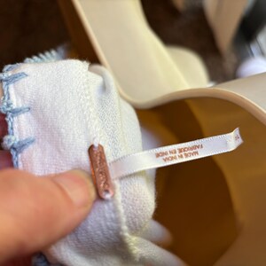 May include: Close-up of a white fabric with light blue stitching along the edge. A small copper-colored tag is attached to the fabric. A white tag with text is also attached to the fabric.