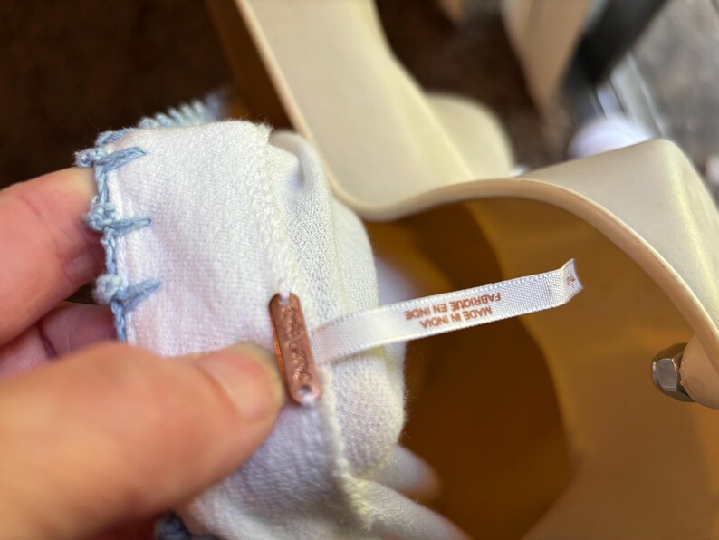 May include: Close-up of a white fabric with light blue stitching along the edge. A small copper-colored tag is attached to the fabric. A white tag with text is also attached to the fabric.