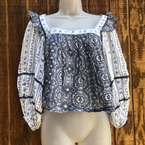 Puede incluir: Una blusa estampada en blanco y azul con mangas largas. La blusa presenta un escote cuadrado con ribete de volantes y un dise&ntilde;o geom&eacute;trico en el corpi&ntilde;o. Las mangas tienen un estampado de rayas florales y est&aacute;n ribeteadas con cinta azul marino.