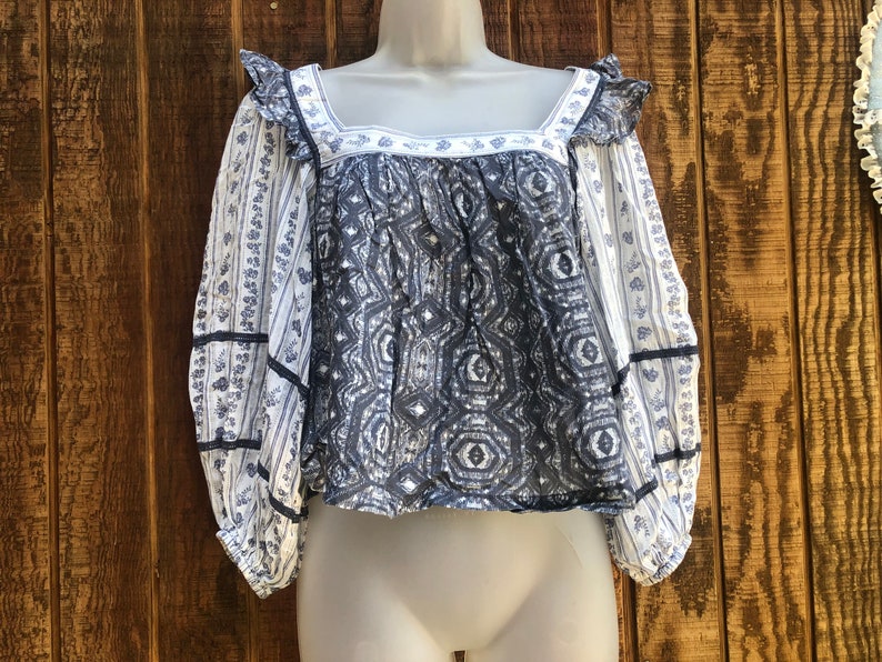 Puede incluir: Una blusa estampada en blanco y azul con mangas largas. La blusa presenta un escote cuadrado con ribete de volantes y un dise&ntilde;o geom&eacute;trico en el corpi&ntilde;o. Las mangas tienen un estampado de rayas florales y est&aacute;n ribeteadas con cinta azul marino.