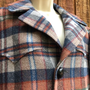 May include: A close-up of a plaid jacket in shades of blue, orange, brown, and gray. The jacket has a notched collar, two front pockets, and a large black button. The fabric appears to be a wool blend.