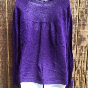 May include: A long-sleeved, purple sweater with a scoop neckline. The sweater is made of a lightweight, sheer material. The garment is displayed on a white mannequin against a wooden background.