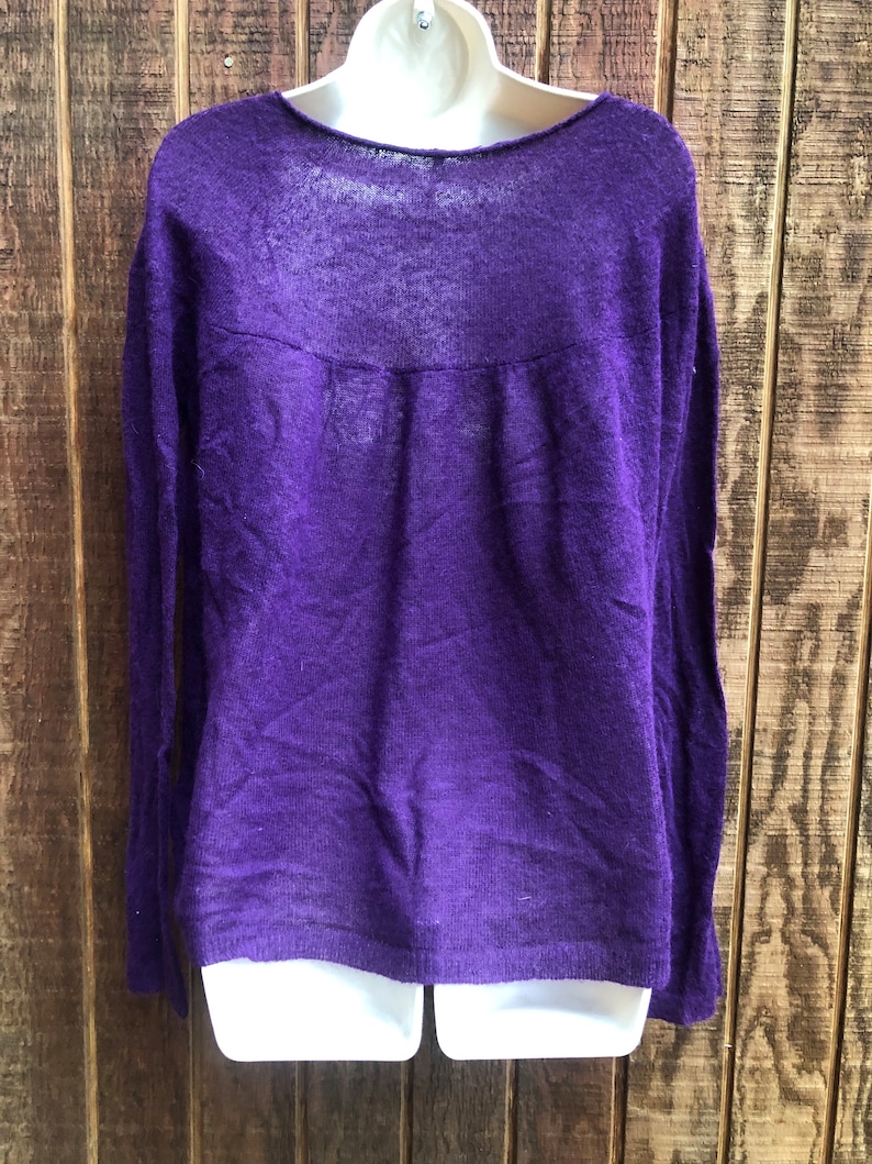 May include: A long-sleeved, purple sweater with a scoop neckline. The sweater is made of a lightweight, sheer material. The garment is displayed on a white mannequin against a wooden background.