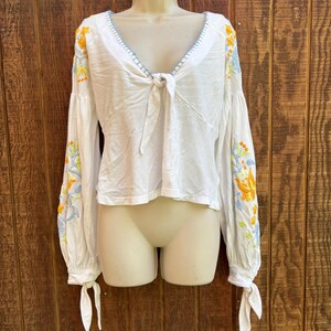May include: White long-sleeve blouse with floral embroidery in yellow, orange, and blue on the sleeves. The blouse features a V-neckline with blue stitching and a tie closure at the front. The sleeves have tie details at the wrists.