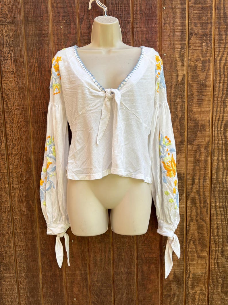 May include: White long-sleeve blouse with floral embroidery in yellow, orange, and blue on the sleeves. The blouse features a V-neckline with blue stitching and a tie closure at the front. The sleeves have tie details at the wrists.