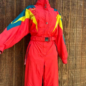 May include: A red FILA ski suit with yellow and teal accents. The one-piece suit has a high collar, a belt, and a zip closure. The suit is made of a shiny material and appears to be vintage.
