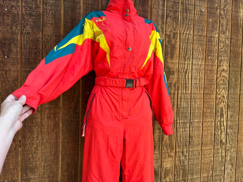 May include: A red FILA ski suit with yellow and teal accents. The one-piece suit has a high collar, a belt, and a zip closure. The suit is made of a shiny material and appears to be vintage.