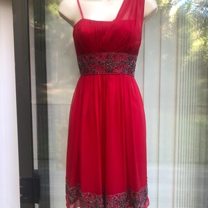 K&ouml;nnte beinhalten: Rotes One-Shoulder-Kleid mit Perlenbesatz an Taille und Saum. Das Kleid wird auf einer Schaufensterpuppe pr&auml;sentiert. Es hat einen flie&szlig;enden Rock und ein eng anliegendes Oberteil. Aus leichtem Stoff.