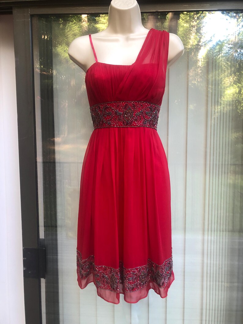 K&ouml;nnte beinhalten: Rotes One-Shoulder-Kleid mit Perlenbesatz an Taille und Saum. Das Kleid wird auf einer Schaufensterpuppe pr&auml;sentiert. Es hat einen flie&szlig;enden Rock und ein eng anliegendes Oberteil. Aus leichtem Stoff.