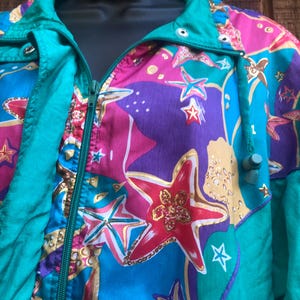 May include: A colorful jacket with a vibrant pattern of stars and shapes in shades of blue, purple, pink, and gold. The jacket has a teal green collar, zipper, and trim. The jacket is a vintage style.