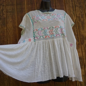 May include: White, embroidered peasant top with colorful geometric patterns across the chest. The top features short sleeves and a flared, gathered hem. The embroidery includes shades of pink, green, blue, and orange. The top is displayed on a black mannequin head.