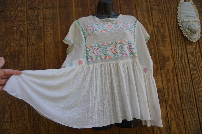 May include: White, embroidered peasant top with colorful geometric patterns across the chest. The top features short sleeves and a flared, gathered hem. The embroidery includes shades of pink, green, blue, and orange. The top is displayed on a black mannequin head.