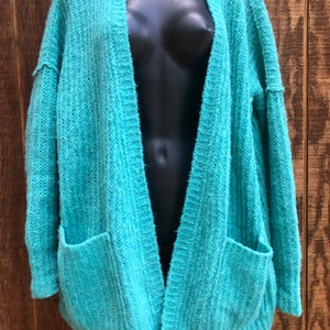 May include: A teal blue open-front cardigan with long sleeves and two patch pockets. The cardigan is made of a soft, fuzzy knit fabric.